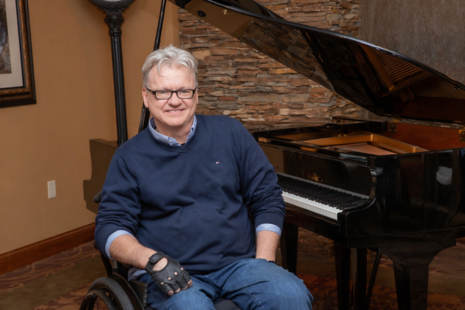 A smiling man in a wheelchair sitting next to a grand piano.