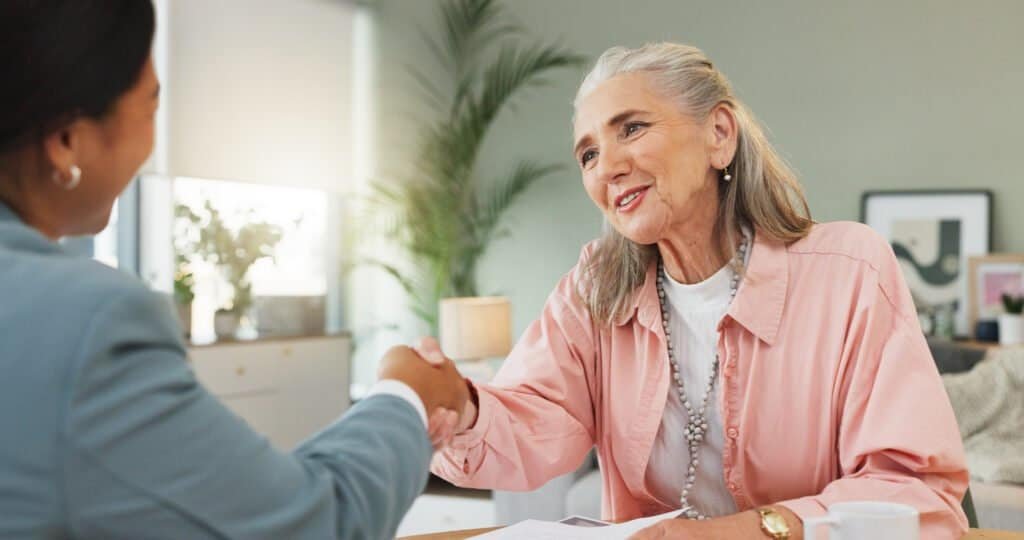 Elderly woman smiling and shaking hands with a businesswoman in an office.