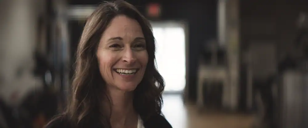Smiling woman in an indoor setting with a supported walk station in the background.